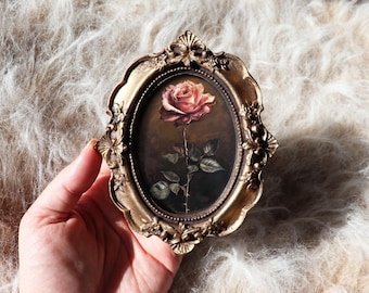 May include: An oval-shaped picture frame with an ornate, metallic gold finish featuring intricate floral and scrollwork details. The frame contains a dark-toned painting of a single, detailed pink rose with green leaves and a thorny stem.