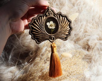 May include: Ornate, antique-style pendant with a pressed white flower encased in a glass-covered frame. The frame is bronze-colored with a decorative tassel hanging below. The pendant is held up by a hand.