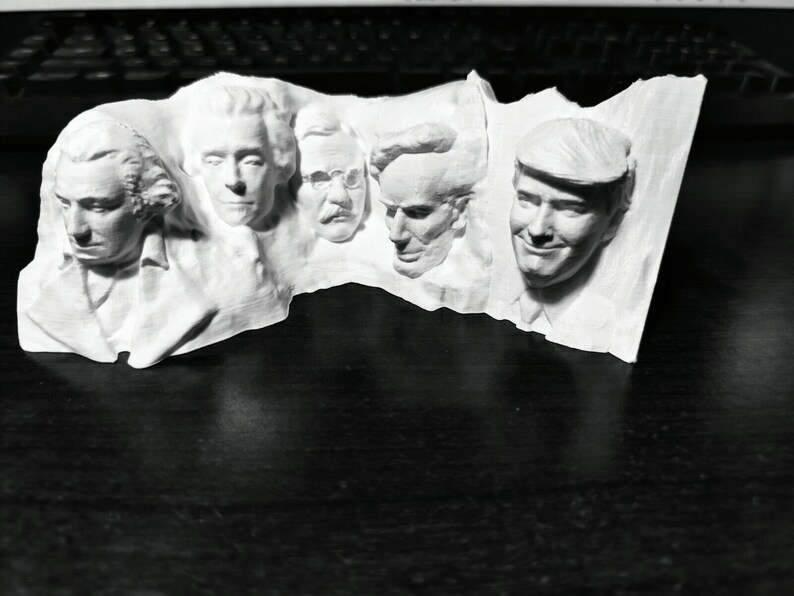 Op de afbeelding: Een witte sculptuur met de gezichten van vier mannen, elk met duidelijke gelaatstrekken. De sculptuur lijkt een gestileerde weergave van een monument te zijn, mogelijk met een historisch of politiek thema.