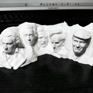 Op de afbeelding: Een witte 3D-geprinte sculptuur met de gezichten van verschillende Amerikaanse presidenten. De sculptuur heeft gedetailleerde gelaatstrekken en is waarschijnlijk een decoratief item of een uniek kunstwerk. De presidenten zijn in een rij gerangschikt, waarbij elk gezicht duidelijk zichtbaar is.