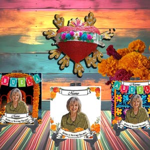 May include: A Day of the Dead themed display featuring three photo frames with a woman's portrait, and a decorative heart. The frames have colorful floral and banner designs with the word "Abuela".