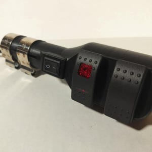 May include: A black device with a silver metal band and a black switch. The device has two black panels with small, round, raised dots. One panel has a red light. A black wire extends from the device.