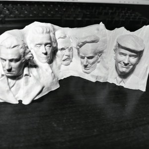 Op de afbeelding: Een witte 3D-geprinte sculptuur met vijf gezichten in reli&euml;f. De gezichten staan naast elkaar en tonen gedetailleerde gelaatstrekken en kapsels. De sculptuur is waarschijnlijk een decoratief item of een uniek kunstwerk.