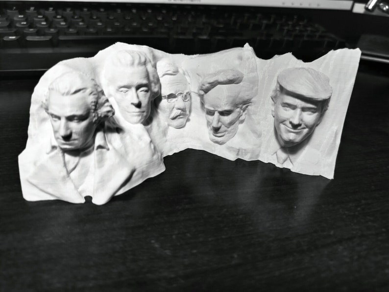 Op de afbeelding: Een witte 3D-geprinte sculptuur met vijf gezichten in reli&euml;f. De gezichten staan naast elkaar en tonen gedetailleerde gelaatstrekken en kapsels. De sculptuur is waarschijnlijk een decoratief item of een uniek kunstwerk.