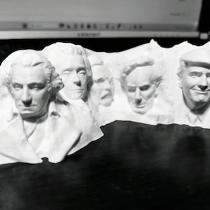 Op de afbeelding: Een witte sculptuur die een berg afbeeldt met de gezichten van verschillende historische figuren erin gebeeldhouwd. De sculptuur bevat de gezichten van George Washington, Thomas Jefferson, Abraham Lincoln, Theodore Roosevelt en Donald Trump.