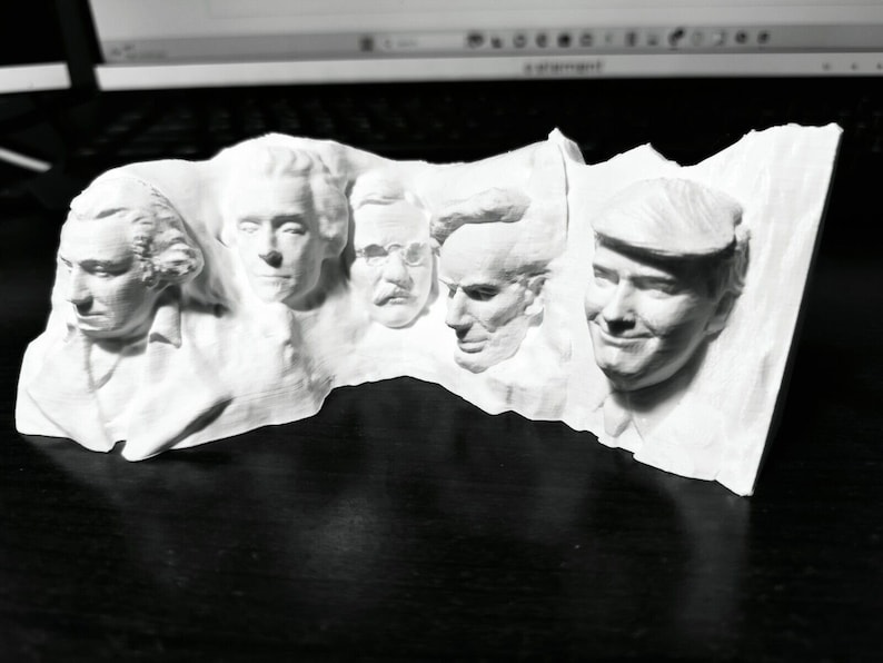 Op de afbeelding: Een witte sculptuur met de gezichten van vijf personen, mogelijk historische figuren, gebeeldhouwd in een rechthoekig blok. De gezichten hebben gedetailleerde kenmerken en kapsels. De sculptuur staat op een donkere ondergrond.