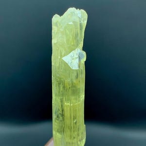 Könnte beinhalten: Ein durchscheinender, blassgelber, länglicher Mineralkristall mit vertikalen Streifen und einer gezackten, unebenen Oberkante. Der Kristall ist vertikal auf einem kleinen, hellen, runden Sockel montiert, der auf einem quadratischen Acryl-Displayblock befestigt ist.