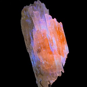 Könnte beinhalten: Ein Cluster aus durchscheinenden, hellorangen Kristallen mit einem blau-violetten Leuchten unter UV-Licht. Die Kristalle sind in einem vertikalen Stapel angeordnet, der an einen Turm erinnert.