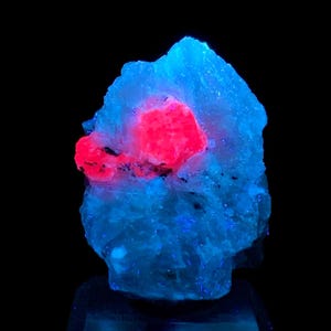 May include: A large, rough, blue crystal with a bright red crystal embedded in the center. The crystal is illuminated with a UV light, causing it to glow.