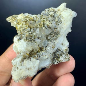 May include: A close-up of a raw mineral specimen. The stone is primarily white with crystalline structures and inclusions of green and black minerals. The rough texture and natural formation are clearly visible.