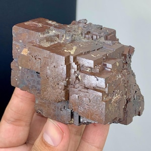 May include: A brown mineral specimen with a cubic crystal structure. The surface has a metallic luster and a rich, earthy color. The mineral appears to be composed of multiple stacked cubes, creating a unique, textured appearance.