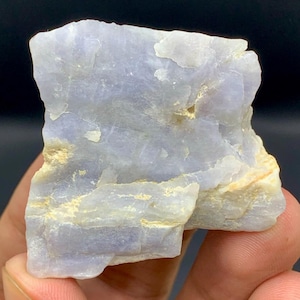 May include: A raw, translucent light blue mineral specimen with a jagged, crystalline structure. The stone features uneven, layered surfaces with subtle white and pale yellow inclusions embedded within the crystalline, light blue rock formation.