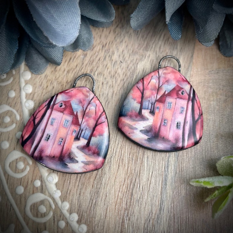 May include: A pair of triangular earrings featuring a watercolour painting of a house in a woodland setting. The house is painted in shades of pink and yellow, and the woodland is painted in shades of red, orange, and green. The earrings are made of polymer clay and have a silver hook.