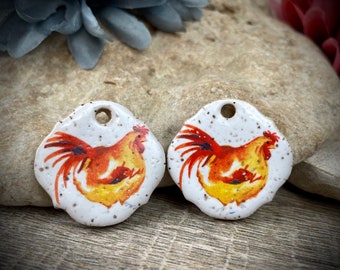 Puede incluir: Dos colgantes de cerámica circulares con un borde irregular y festoneado. La superficie es blanca con pequeñas motas marrones y presenta una colorida ilustración de un gallo con plumas naranjas, amarillas y rojas. Cada colgante tiene un orificio.