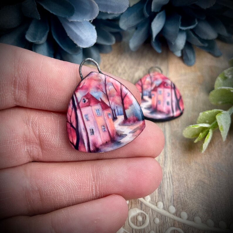 May include: A pair of triangular ceramic earrings featuring a painted image of a house in a woodland setting. The house is painted in shades of pink and yellow, with a red roof. The woodland is painted in shades of green and brown. The earrings are suspended from silver jump rings.