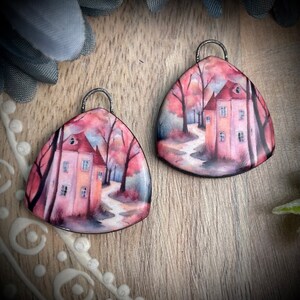 May include: A pair of triangular earrings featuring a watercolour painting of a house in a woodland scene. The house is painted in shades of pink and orange, with a red roof. The trees are painted in shades of green and brown. The earrings are made of metal and have a silver finish.