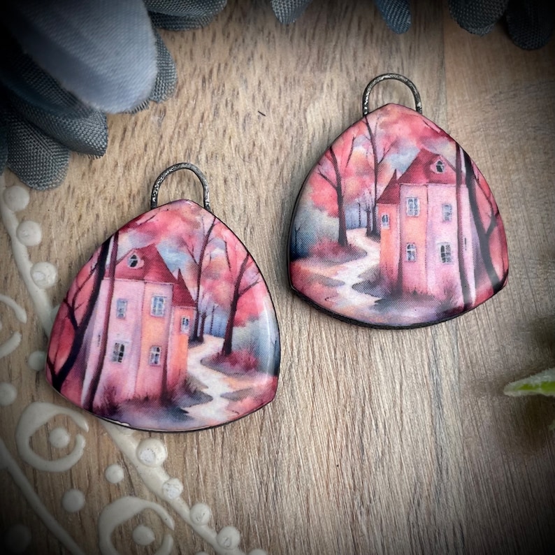 May include: A pair of triangular earrings featuring a watercolour painting of a house in a woodland scene. The house is painted in shades of pink and orange, with a red roof. The trees are painted in shades of green and brown. The earrings are made of metal and have a silver finish.