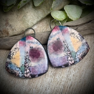 Puede incluir: Un par de pendientes geométricos abstractos con forma de triángulo. Los pendientes están hechos de arcilla polimérica y presentan un diseño colorido con tonos de morado, rosa, amarillo y azul. Los pendientes tienen un contorno negro y están unidos a ganchos de oreja plateados.