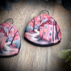 May include: A pair of triangular earrings featuring a watercolour painting of a house in a woodland setting. The house is pink with a red roof and white detailing. The trees are a mix of brown and green. The background is a soft pink and blue.