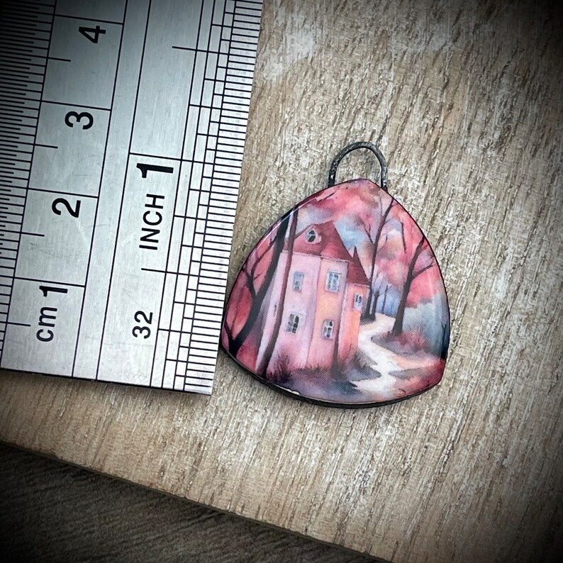 May include: A triangular pendant featuring a painted image of a pink house with a path leading to it. The house is surrounded by trees and the background is a pink and blue sky.