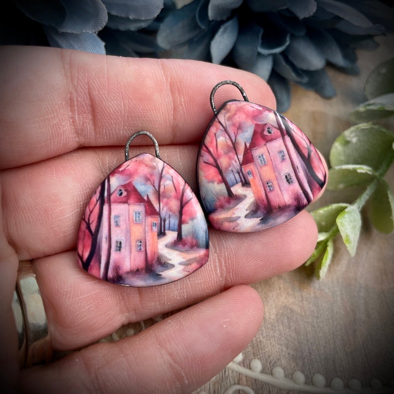 May include: A pair of pink and white triangle-shaped earrings with a painted image of a house in a forest. The earrings have a silver-coloured metal loop for hanging.