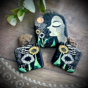 May include: Three square pendants featuring black backgrounds with white and yellow floral designs. One pendant displays a profile of a face with closed eyes, a sun, and flowers. The other two pendants show white and yellow flowers with swirling patterns.