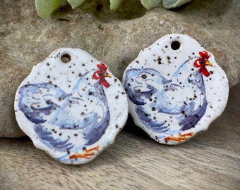 Puede incluir: Dos adornos de cerámica blanca en forma de pollo con motas marrones y detalles pintados en azul y blanco. Los pollos están mirando hacia la derecha y tienen crestas rojas.
