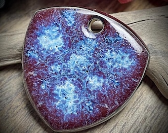Puede incluir: Un colgante de cerámica de color morado oscuro y azul con un patrón de remolino blanco y un pequeño agujero para colgar.