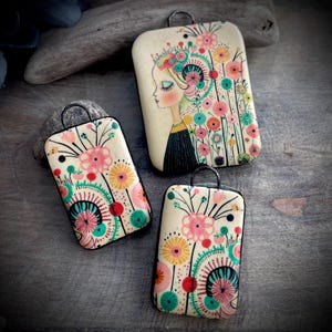 May include: Three rectangular pendants with rounded corners and black metal bails. The cream-colored surfaces feature illustrations of stylized flowers in shades of pink, yellow, teal, and red. One pendant depicts a profile of a person with floral hair.