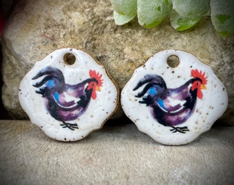 Puede incluir: Dos amuletos de cerámica con forma de gallo con fondo blanco y un gallo morado con cresta y barbilla rojas. Los amuletos tienen un agujero en la parte superior para colgar.