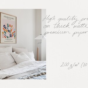 May include: A white bedroom with a framed print on the wall. The print features a pink background with the word "FEMINISME" and a design of colorful gender symbols. The text "LA REVOLUTION" is also included on the print. The bed has white linens and pillows. A lamp is on a wooden stand next to the bed. The text "High quality print on thick matte premium paper 200 g/m&sup2; (80 lb)" is written in cursive on a white background.