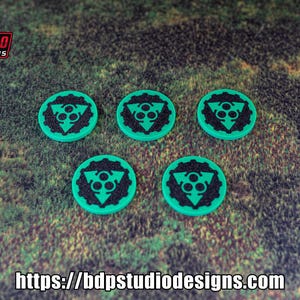 May include: Five teal green round tokens with a black symbol of three circles inside a triangle. The tokens are on a green grassy surface.