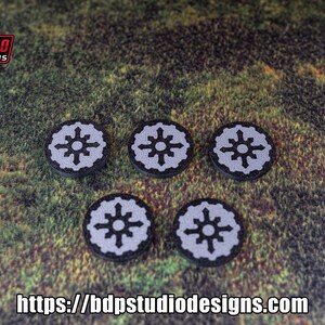 May include: Five black and white circular tokens with a stylized starburst design. The tokens are on a green and brown textured surface.