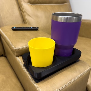 May include: A black cup holder with two slots for cups. A yellow cup and a purple tumbler are in the slots. The cup holder is on a brown leather couch.