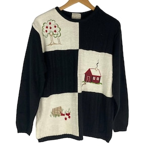 May include: A black and cream-colored sweater with embroidered patches. The patches feature a tree with red apples, a red house, and a basket of apples. The sweater is on a wooden hanger.
