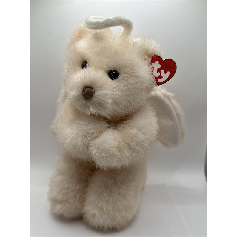 May include: A cream-coloured plush dog-like toy with a halo and wings. It has a brown nose, black eyes, and a red heart-shaped tag. The soft toy is in a sitting position.