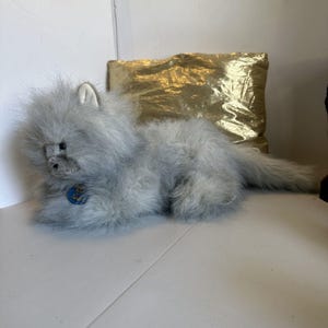 May include: A plush, gray cat toy with a fluffy coat and a small blue and gold emblem on its chest. The cat is lying down, with a long tail extending to the right. A gold pillow is in the background.
