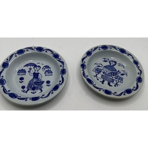 May include: Two small, white ceramic plates with a blue floral border and a central illustration. One plate depicts a woman with an umbrella, the other a woman with a cart. The plates are likely vintage and suitable for serving small portions.
