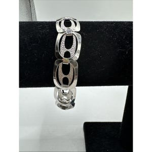 Op de afbeelding: Zilverkleurige armband met een open ontwerp. De armband heeft een reeks onderling verbonden, langwerpige ovale schakels. Elke schakel heeft een centraal, getextureerd ovaal element. De armband wordt gepresenteerd tegen een zwarte achtergrond.