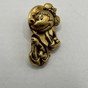 Könnte beinhalten: Ein goldfarbener Ansteckpin mit einer stilisierten Darstellung einer Cartoon-Mausfigur. Der Pin hat ein detailliertes Design, wobei die Maus einen Hut trägt und einen Stab hält. Der Pin ist wahrscheinlich aus Metall und hat eine klassische, Vintage-Ästhetik.