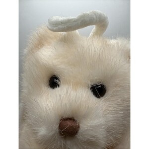 May include: A close-up shot of a cream-coloured plush bunny with a fluffy texture. The bunny has black eyes and a small, brown nose. A white, curved piece extends from the top of its head, resembling a halo. The image highlights the soft, furry details of the toy.