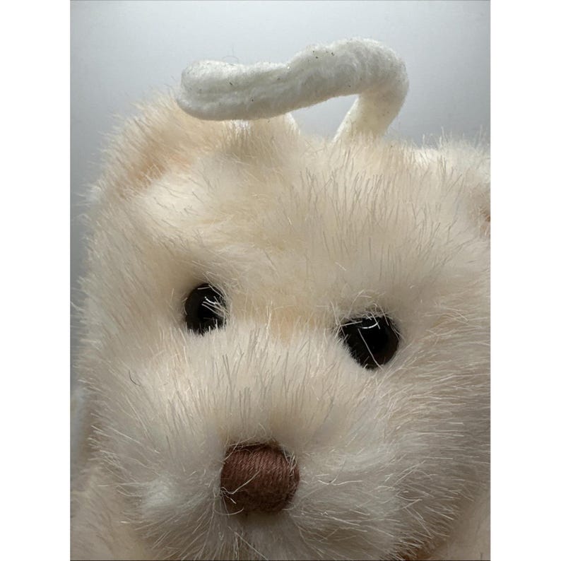 May include: A close-up shot of a cream-coloured plush bunny with a fluffy texture. The bunny has black eyes and a small, brown nose. A white, curved piece extends from the top of its head, resembling a halo. The image highlights the soft, furry details of the toy.