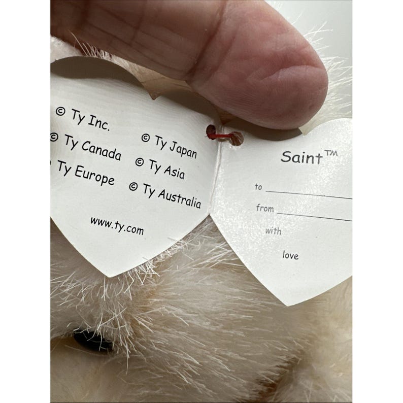 May include: Close-up of a plush toy with a heart-shaped tag. The tag reads "Saint" and includes the text "to", "from", and "with love". The tag also includes the text "&copy; Ty Inc.", "&copy; Ty Canada", "&copy; Ty Europe", "&copy; Ty Japan", "&copy; Ty Asia", and "&copy; Ty Australia".