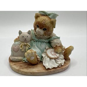 Peut inclure: Figurine d'un chaton assis, vêtu d'une robe et d'un nœud bleu clair, tenant un napperon blanc avec un cœur. Une figurine de chat plus petite, blanche, avec un cœur est assise à côté du chaton. Sur une base imitation bois.