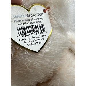May include: A soft, white plush toy with a heart-shaped tag. The tag states "SAFETY PRECAUTION" and includes a barcode and instructions: "Retain Tag For Reference. For Ages 3 And Up Surface Wash."