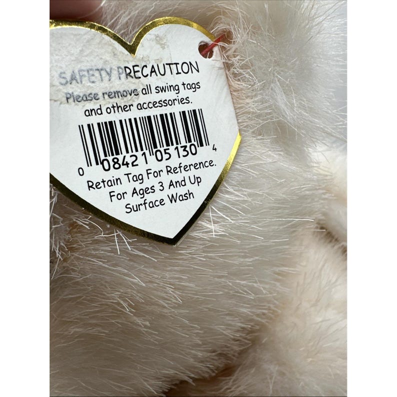 May include: A soft, white plush toy with a heart-shaped tag. The tag states "SAFETY PRECAUTION" and includes a barcode and instructions: "Retain Tag For Reference. For Ages 3 And Up Surface Wash."