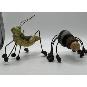 Könnte beinhalten: Zwei kleine Figuren von Insekten. Ein grüner Grashüpfer mit braunen und weißen Markierungen und eine schwarz-weiße Spinne mit einem braunen Kopf.