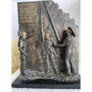 May include: A bronze sculpture of three firefighters raising an American flag. The sculpture is set on a black base and includes the quote "Greater love has no man than this, that he lay down his life for his friends." John 15:13.
