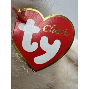 May include: A close-up of a red heart-shaped tag with gold edging, displaying the word "Classic" and the "ty" logo in white. The tag is attached to a plush toy with white fur.