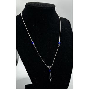 May include: A silver necklace with a delicate chain and a pendant. The necklace features three small blue beads and a teardrop-shaped pendant with a dark blue stone. The necklace is displayed on a black mannequin.